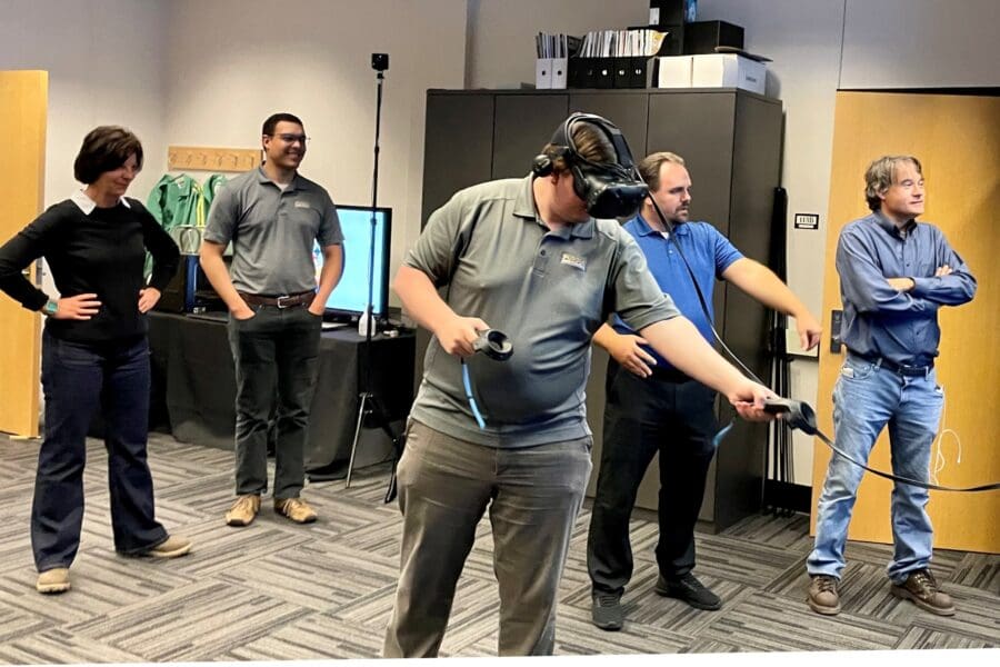 Visitor participates in hands-on experiences of AR & VR projects in the visualization lab. Participant is standing while wearing a tethered VR headset and manipulating had held controllers surrounded by several people observing