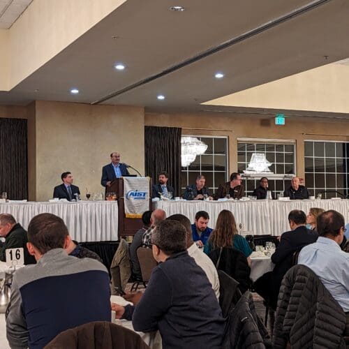 Several round tables with people seated in chairs. Long table in the front of the room with man at podium