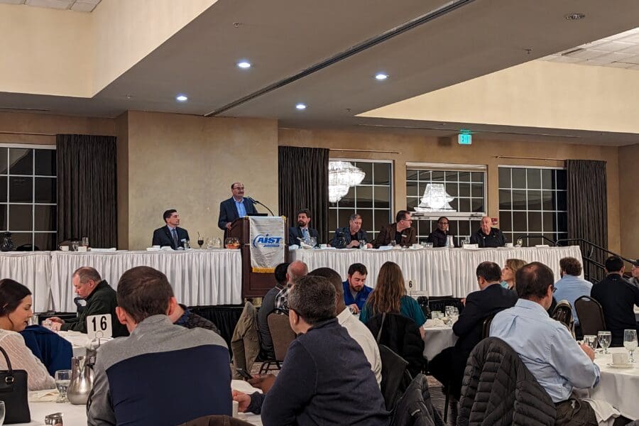 Several round tables with people seated in chairs. Long table in the front of the room with man at podium