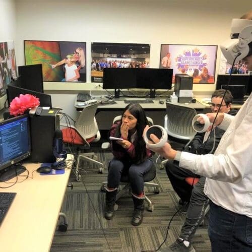 Visitor participates in hands-on experiences of AR & VR projects in the visualization lab. Participant is standing while wearing a tethered VR headset and manipulating hand held controllers surrounded by student researchers observing