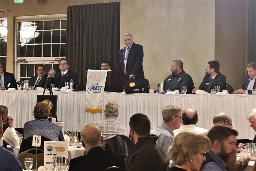 Several round tables with people seated in chairs. Long table in the front of the room with man at podium