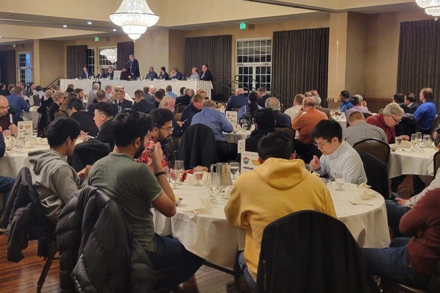 Several round tables with people seated in chairs. Long table in the front of the room with man at podium