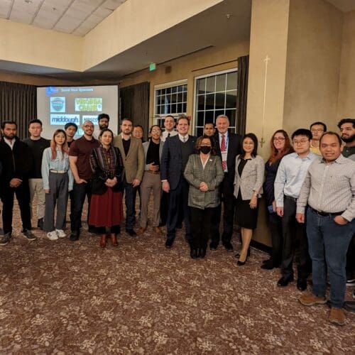 A large group of individuals in business professional attire stand together in front of a large screen.