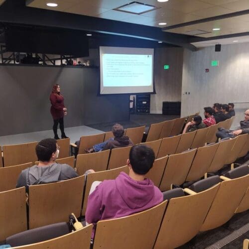 Woman standing in front of screen in auditorium with several people seated in rows of chairs