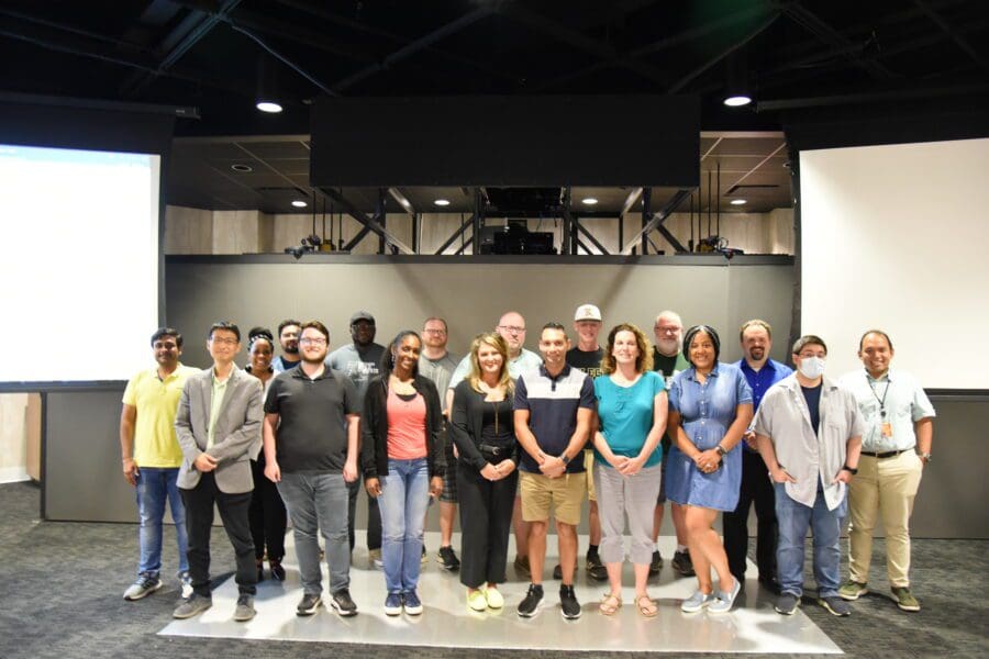 A large group of individuals in business casual attire stand together in front of a large screen.