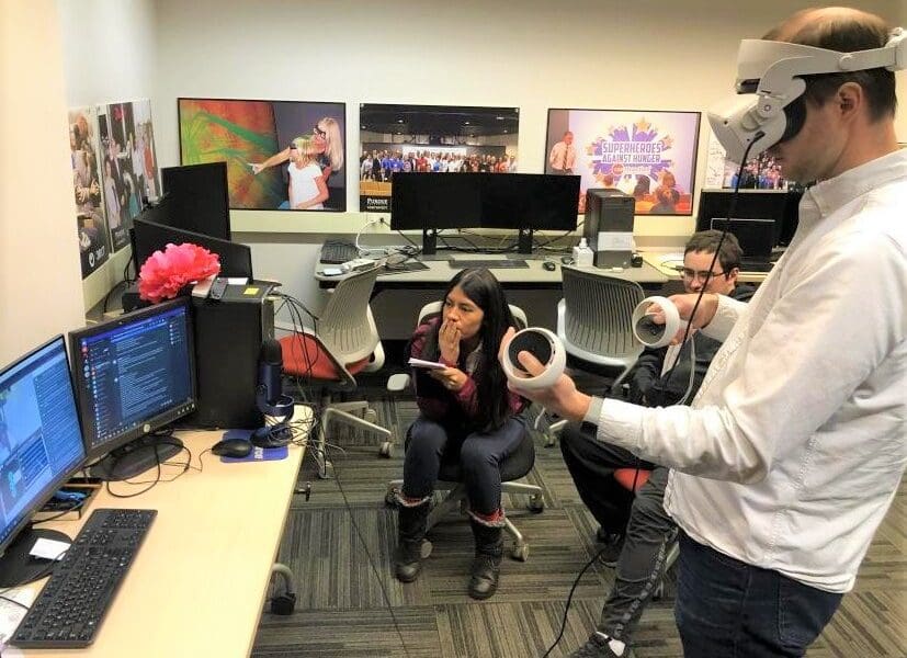 Visitor participates in hands-on experiences of AR & VR projects in the visualization lab. Participant is standing while wearing a tethered VR headset and manipulating hand held controllers surrounded by student researchers observing