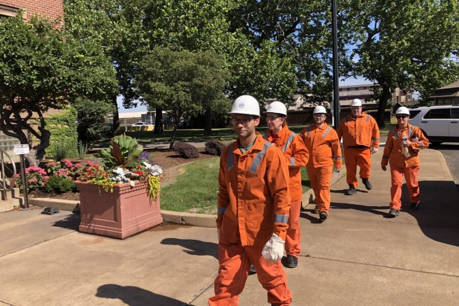 Visitors walking outdoors wearing bright orange uniform jumpsuits and white hard hats and safety goggles