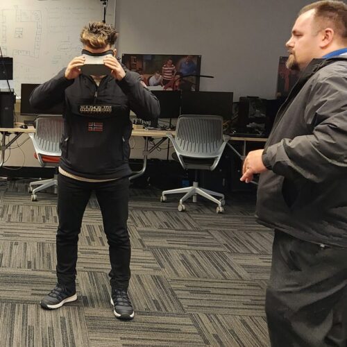 Visitor participates in hands-on experiences of AR & VR projects in the visualization lab. Participant is standing while holding VR goggles. Another individual gives instruction.