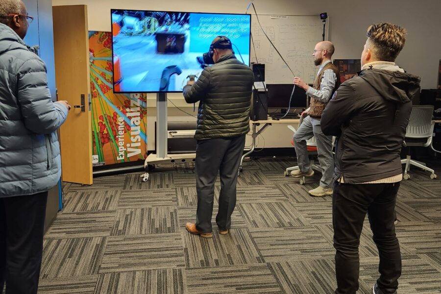 Visitor participates in hands-on experiences of AR & VR projects in the visualization lab. Participant is standing while wearing a tethered VR headset and manipulating hand held controllers surrounded by several people observing