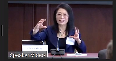 Woman in business attire seated at table with microphone speaking in a conference room