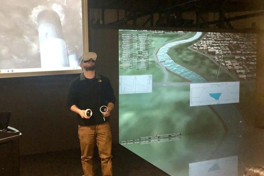 Student participates in hands-on experiences of AR & VR projects in the Immersive Theater at CIVS. Participant is standing while wearing VR headset and manipulating hand held controls. The large screen features what the participant is seeing in the goggles.