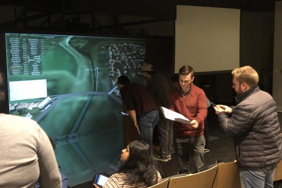 Students participant in virtual flood simulator course in the Immersive Theater at CIVS. The large screen shows the simulation while students talk and take notes.