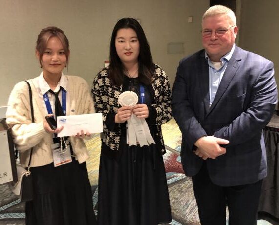 3 individuals in business professional attire stand together, 2 holding awards.