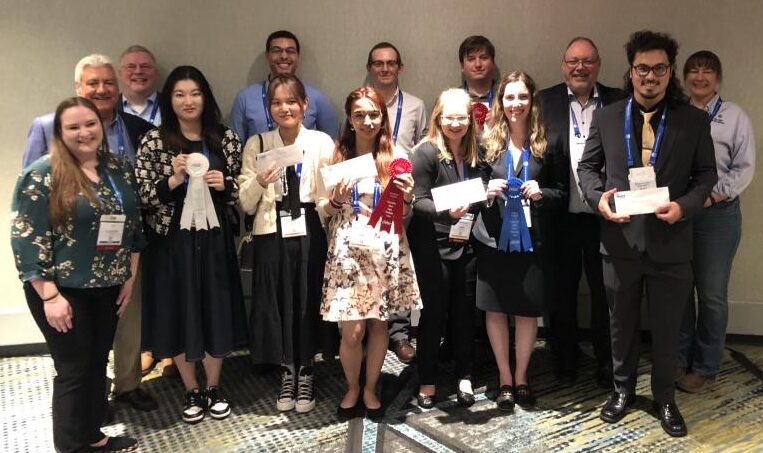 A large group of individuals in business professional attire stand together holding various awards