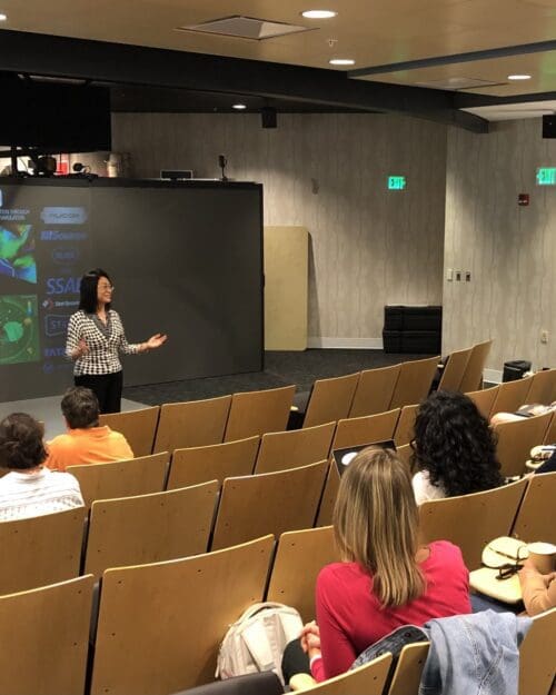 Woman standing in front of screen in auditorium with several people seated in rows of chairs