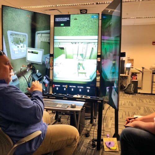 An individual operates a crane training simulator in the CIVS visualization lab while giving instruction to another individual