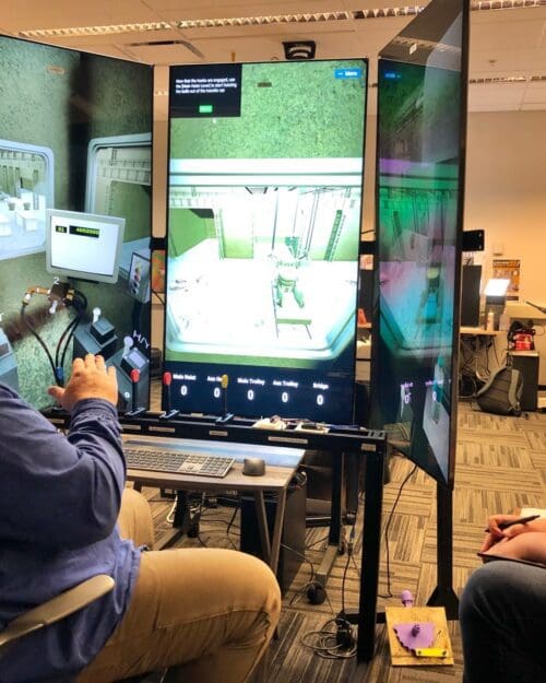 An individual operates a crane training simulator in the CIVS visualization lab while giving instruction to another individual