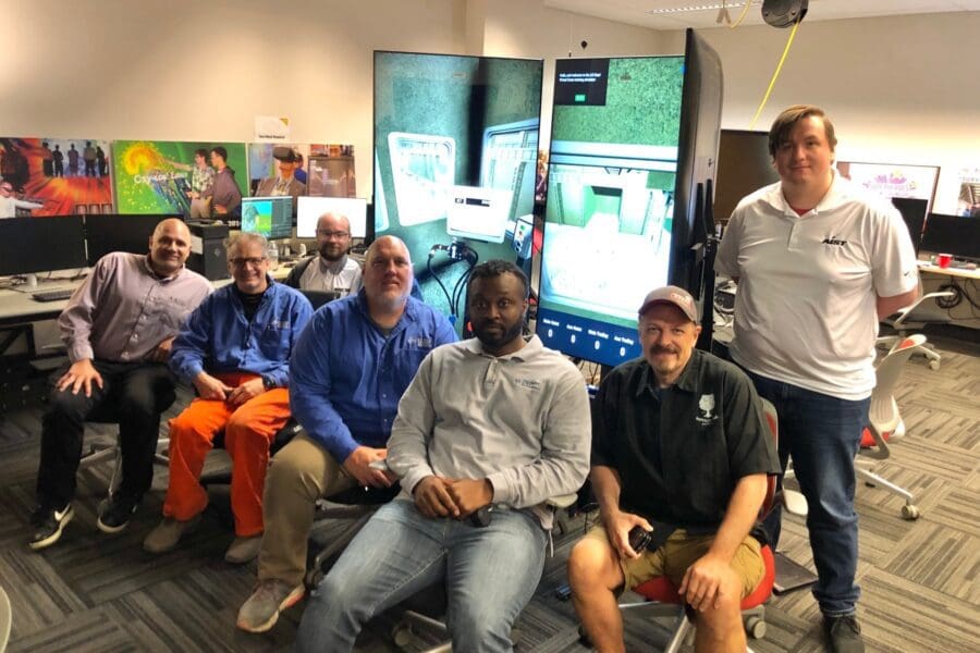 A group of individuals standing in a row in the CIVS Visualization lab. Behind them is the crane simulator module on 3 large screens.