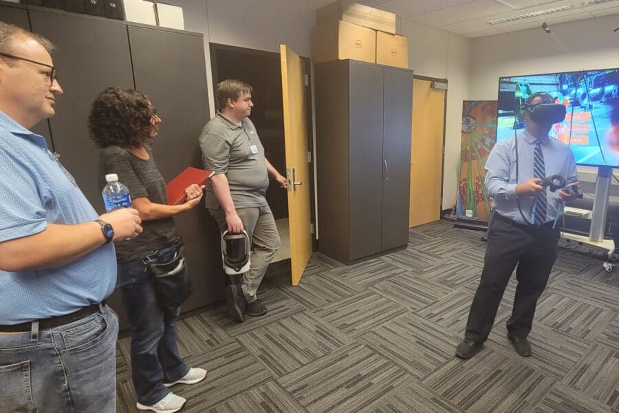 An individual in business professional attire stands wearing a VR headset and operating handheld controls in the CIVS visualization lab while other individuals watch. The screen behind them shows what they see in the VR headset: the fire extinguisher simulator