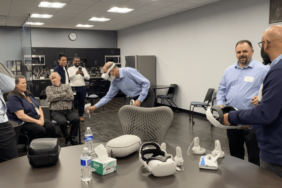 An individual wearing a VR headset and operating handheld controls stands in the middle of a conference room while others watch and talk among themselves