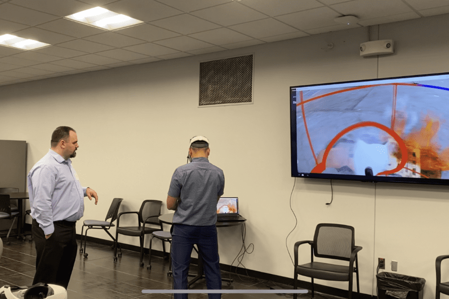 An individual wearing a VR headset and operating handheld controls stands in the middle of a conference room while another individual gives instructions. The screen next to them shows what the individual sees in the VR headset.