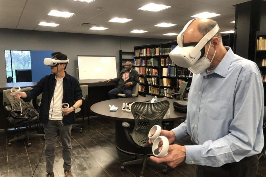 Two individuals wearing a VR headset and operating handheld controls stands in the middle of a conference room while another individual sits and watches