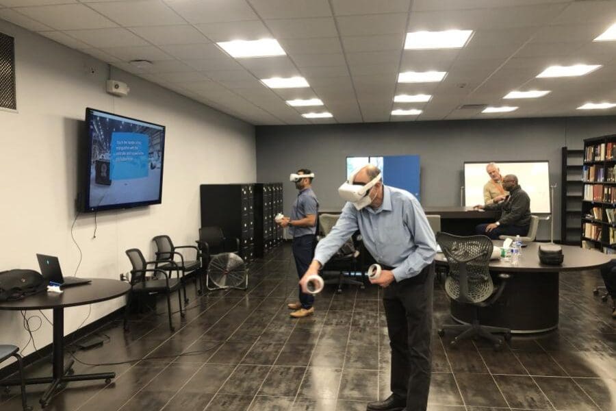 An individual wearing a VR headset and operating handheld controls stands in the middle of a conference room. The screen next to them shows what the individual sees in the VR headset: the fire extinguisher simulator.