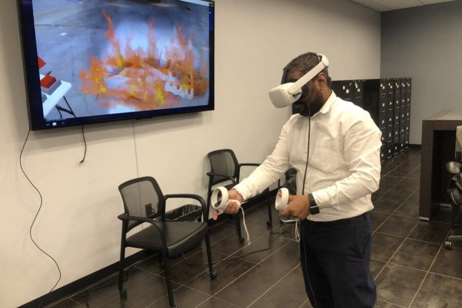 An individual wearing a VR headset and operating handheld controls stands in the middle of a conference room. The screen next to them shows what the individual sees in the VR headset: the fire extinguisher simulator.