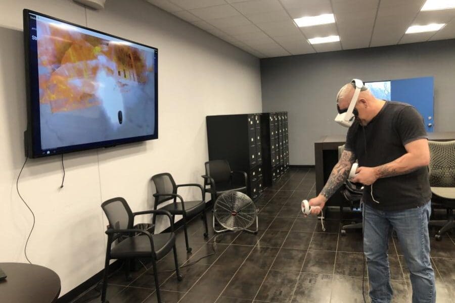 An individual wearing a VR headset and operating handheld controls stands in the middle of a conference room. The screen next to them shows what the individual sees in the VR headset: the fire extinguisher simulator.