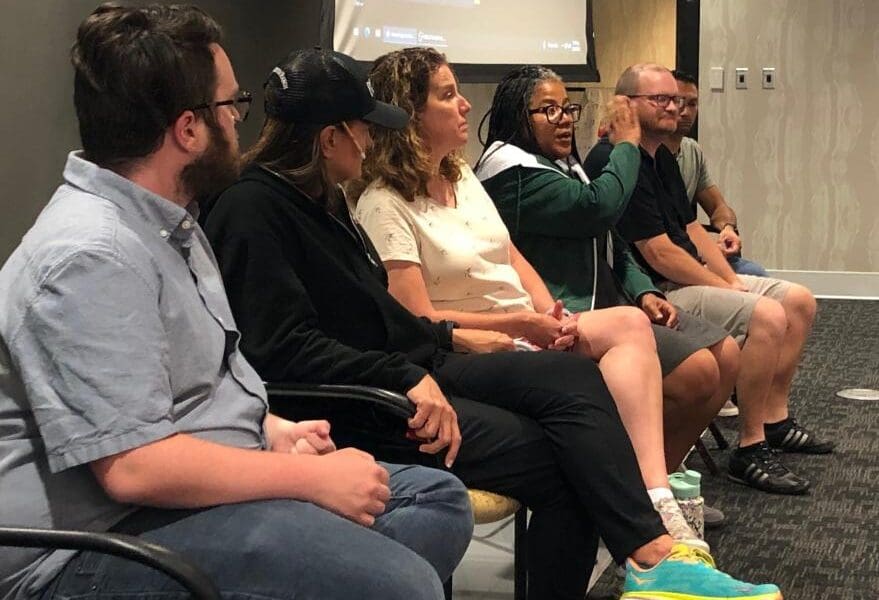 Several people seated in a row at the front of a conference room, a lady speaks while the others face her