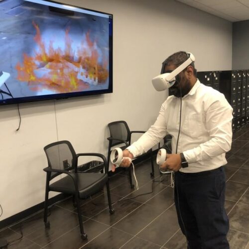 An individual wearing a VR headset and operating handheld controls stands in the middle of a conference room. The screen next to them shows what the individual sees in the VR headset: the fire extinguisher simulator.