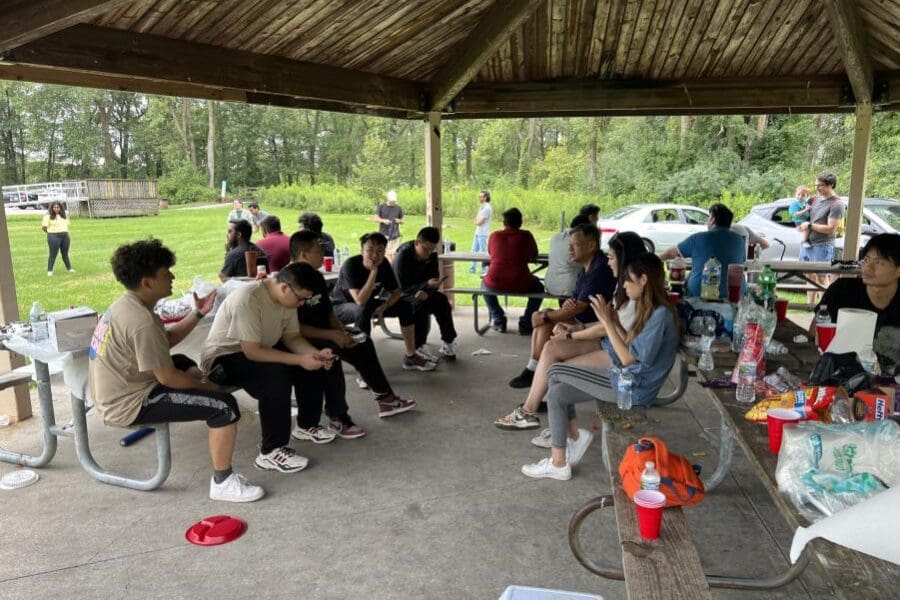 A large group of individuals talking to each other are seated at tables under a canopy at a park