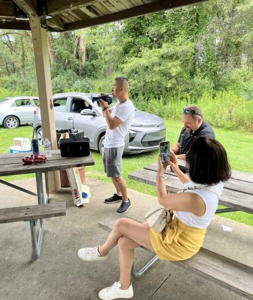 An individual playing the trumpet stands under a canopy in a park while another individual records them on their phone