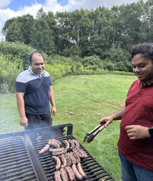 An individual grills sausage on a charcoal grill in a park while another individual stands and watches