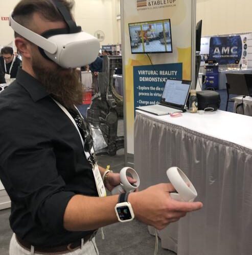 An individual standing in a conference hall wearing a VR headset and operating handheld controls with several booths in the background