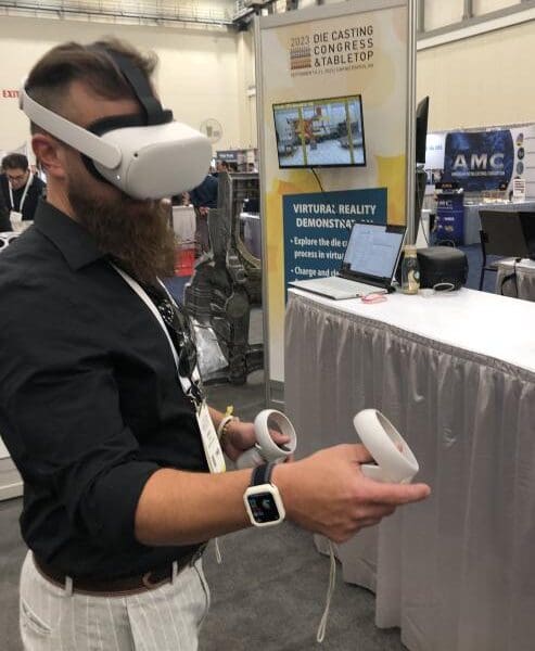 An individual standing in a conference hall wearing a VR headset and operating handheld controls with several booths in the background