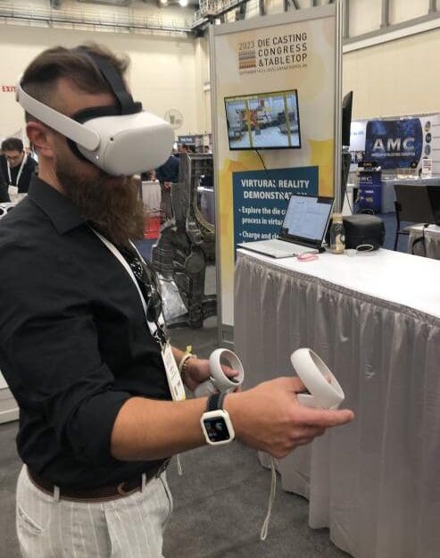 An individual standing in a conference hall wearing a VR headset and operating handheld controls with several booths in the background