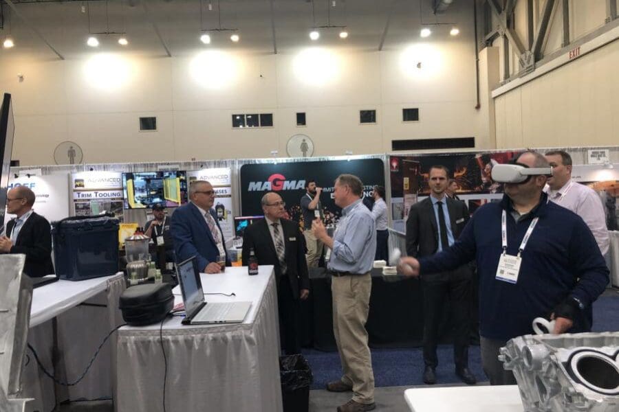 An individual standing in a crowded conference hall wearing a VR headset and operating handheld controls with several individuals and booths in the background