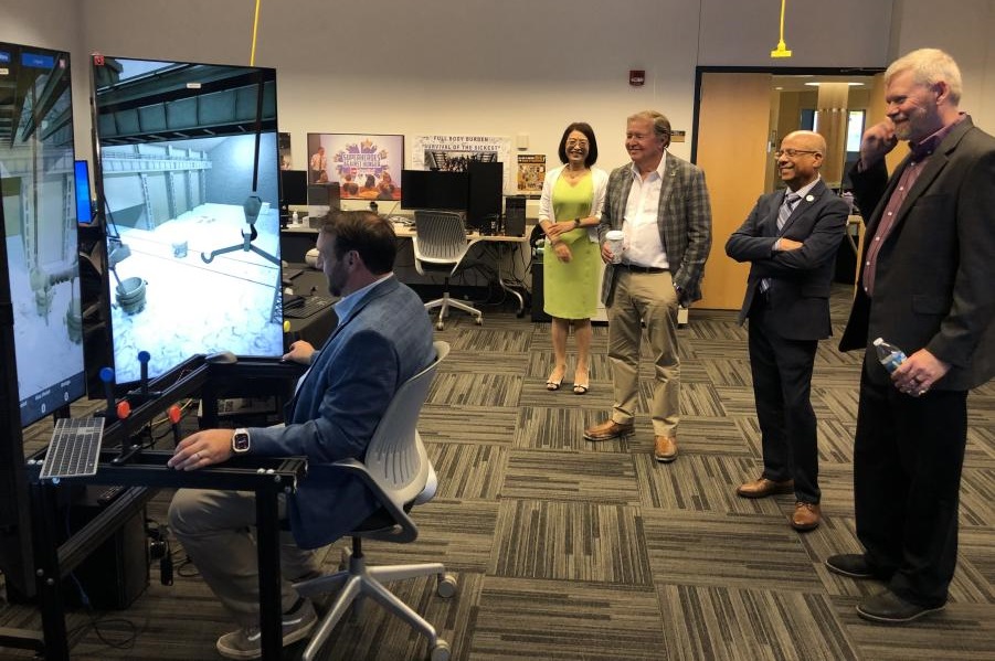 An individual in business professional attire operates a crane training simulator in the CIVS visualization lab while other individuals watch and smile