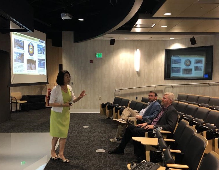 Woman standing in front of screen in auditorium with two people seated in the front row of chairs