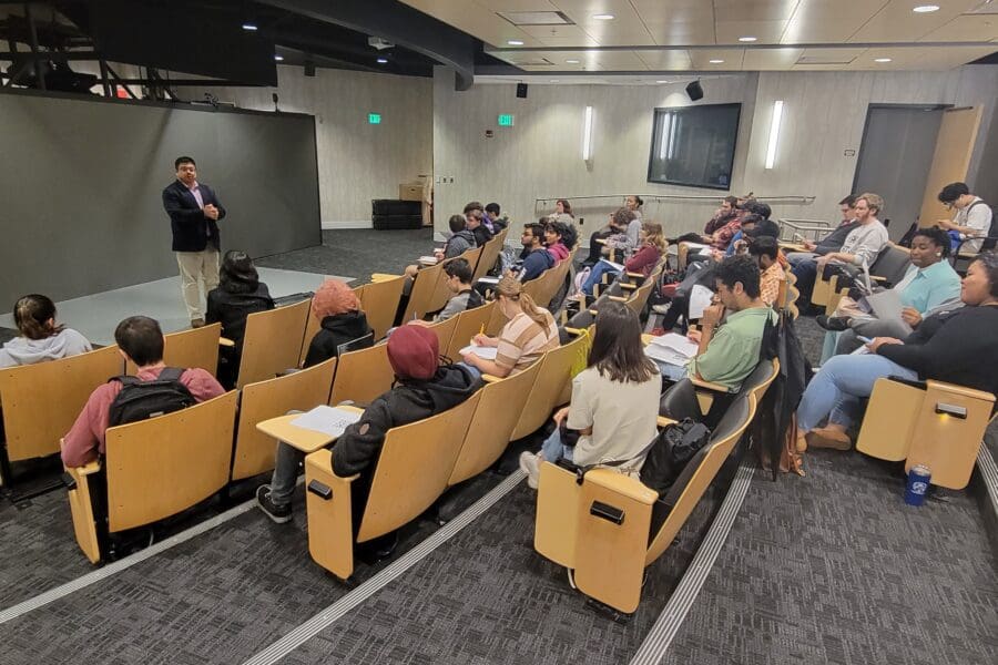 Man standing in front of screen in auditorium with several people seated in rows of chairs