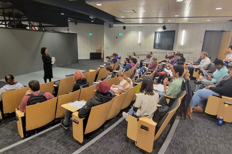 Woman standing in front of screen in auditorium with several people seated in rows of chairs