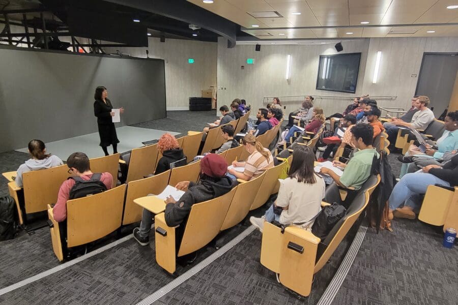 Woman standing in front of screen in auditorium with several people seated in rows of chairs