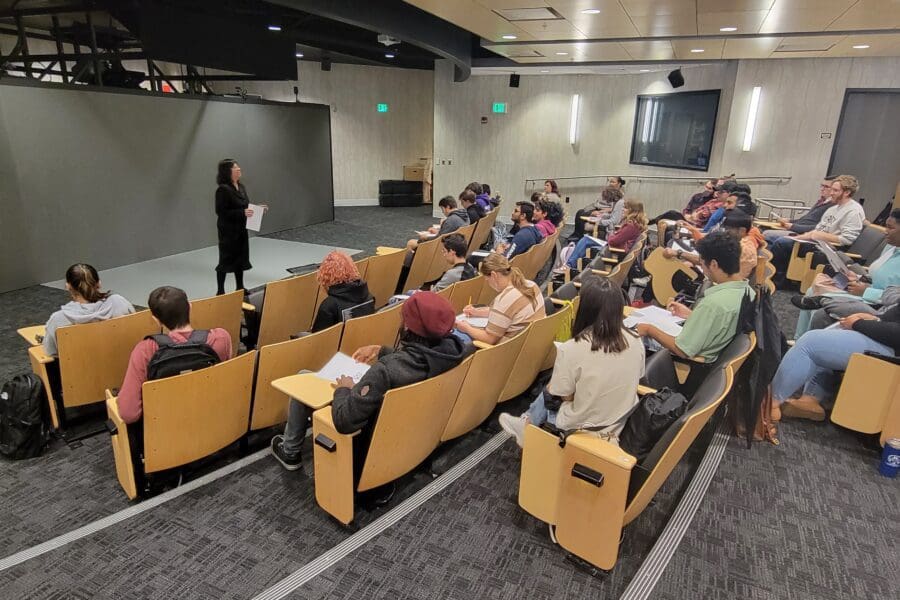 Woman standing in front of screen in auditorium with several people seated in rows of chairs