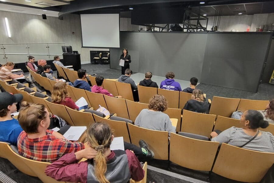 Woman standing in front of screen in auditorium with several people seated in rows of chairs