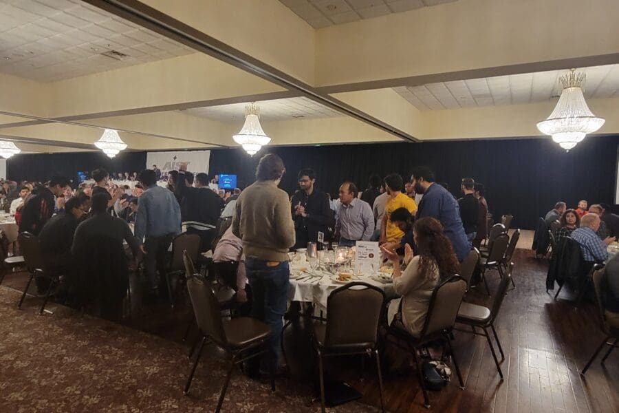 Several round tables with people seated in chairs. Long table in the front of the room with man at podium. Few people are standing in recognition.