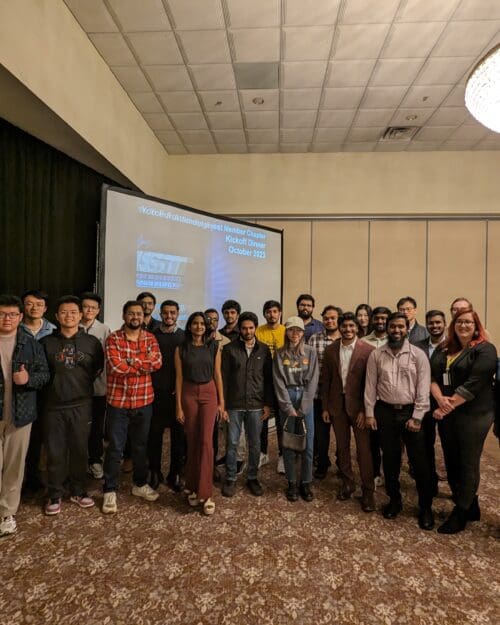 A large group of individuals in business professional attire stand together in front of a large screen.