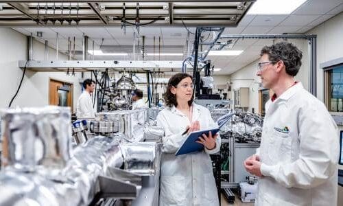 Man and woman in white lab coats standing next to metal tubing in a professional engineering lab setting with several computers, monitors, and wiring