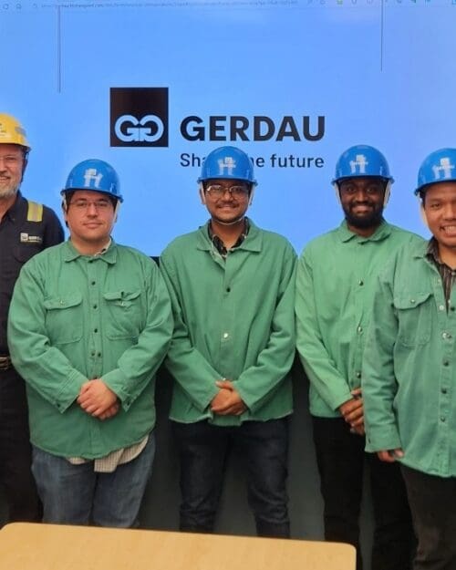 A row of 7 men in uniforms, 4 in all green with blue hard hats and 3 in navy with high vis stripes on the shoulders and arms with yellow hard hats standing in a conference room. Screen in the background contains Gerdau company logo.