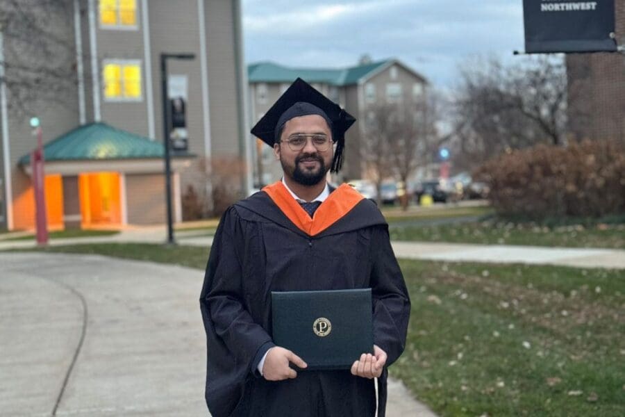 Young student in cap and gown standing in front of academic building holding diploma